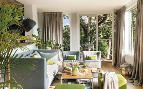 Sofá gris y otomanas verdes crean un ambiente acogedor. Plantas exuberantes y luz natural inundan el salón.