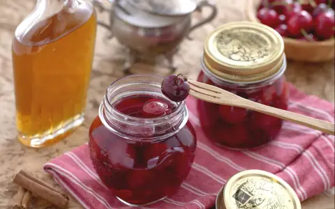 Frascos de mermelada de cerezas casera, una botella de licor y canela en rama. Una cereza en un tenedor de madera lista para probar.