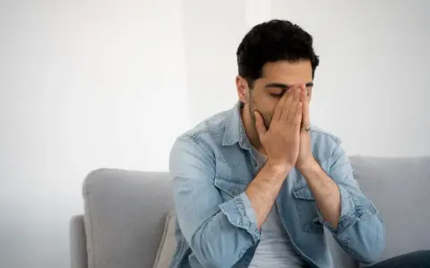 Mann sitzt auf Sofa, Hände vors Gesicht gepresst, Augen geschlossen. Fühlt sich nach dem Rauchen schwindelig.