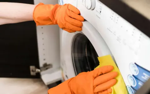 Hände in orangen Handschuhen reinigen die Waschmaschine. Ein gelber Lappen wischt über die Oberfläche, um sie sauber zu halten.