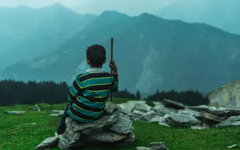 Junge sitzt auf Steinen, blickt in nebelverhangene Berge. Ein Stock in der Hand, wie für einen meditationstext zum vorlesen.