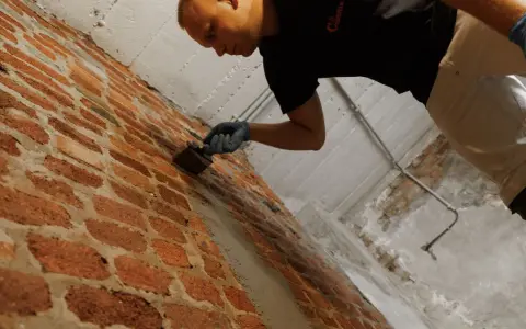 Mann mit Handschuhen trägt eine Masse auf eine Ziegelwand auf, um Anschlussfugen abzudichten.