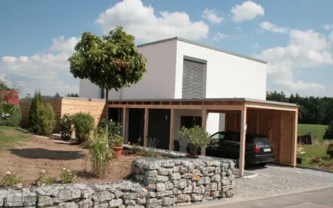 Modernes, weißes Haus mit Holzcarport und Gabionenmauer. Ein Beispiel, wie man ein low budget haus bauen kann, das dennoch stilvoll aussieht.