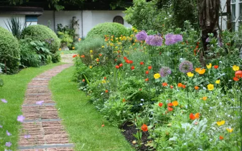 Ein geschwungener Weg aus Ziegelsteinen schlängelt sich durch einen üppigen bauerngarten mit leuchtenden Blumen und grünem Rasen.