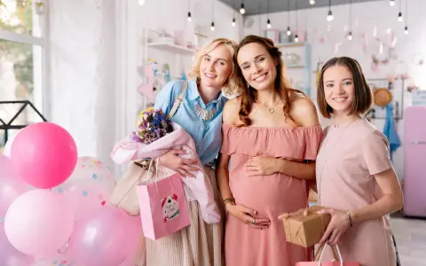Trzy uśmiechnięte kobiety świętują baby shower. Jedna z nich jest w ciąży, a pozostałe dwie trzymają prezenty i balony.