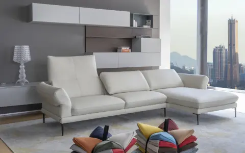 Un divano bianco in pelle, con cuscini colorati sparsi sul tappeto, davanti a una finestra con vista sulla città al tramonto. L'alba calia illumina la scena.