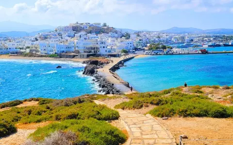 A stone path leads to a causeway connecting to the whitewashed town of Naxos, Greece, with vibrant blue waters lapping at the shore.