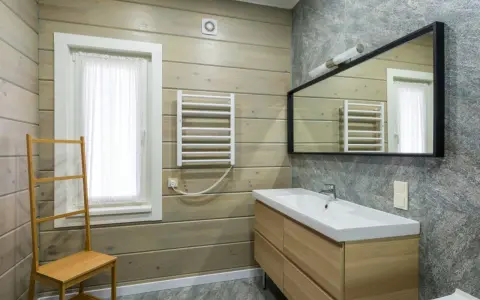 Salle de bain moderne avec murs en bois clair et pierre grise. Un meuble vasque en bois, un miroir, un radiateur et une échelle en bambou pour habiller un mur.