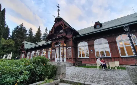 Drewniany pawilon z zegarem i karłowata studzienka przed wejściem. Na zewnątrz siedzą ludzie przy stoliku.