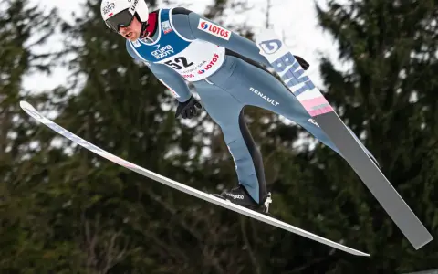 Skoczek narciarski w locie, numer 52, podczas kwalifikacji w skokach. W tle drzewa.