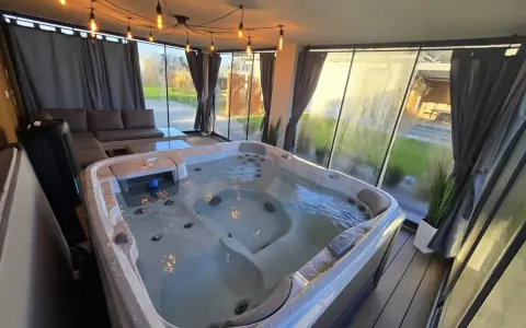 Relaks w jacuzzi pod ciepłym światłem lamp w przeszklonym tarasie zimowym. Obok wygodna sofa i stolik.
