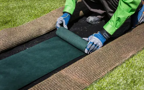 Manos enguantadas desenrollan césped artificial verde sobre una base negra. Paso clave para saber como colocar cesped artificial.