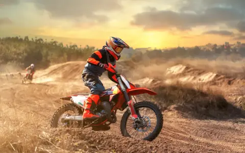 Un pilota di **enduro motocross** sfreccia su un terreno polveroso al tramonto, sollevando terra.
