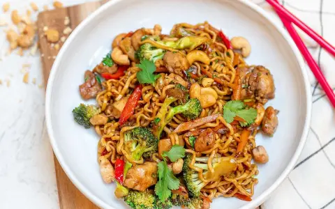 En skål med nudlar, kyckling, broccoli, paprika och cashewnötter. En perfekt allt i ett mat-rätt, redo att ätas med röda ätpinnar.