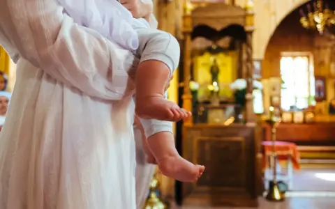 Chrzestna trzyma dziecko w kościele. W tle widać ikonostas i ławki.