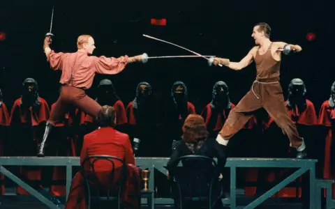 Scen ur Hamlet där två män duellerar med svärd. Bakom dem står figurer i röda dräkter med masker. Vad handlar Hamlet om? Kanske svek och hämnd.