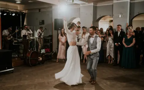 Para młoda tańczy na parkiecie podczas wesela w remizie. W tle zespół gra muzykę, a goście obserwują.