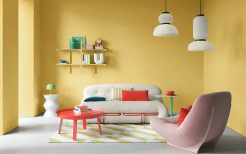 Ein Wohnzimmer mit gelben Wänden und einer auffälligen farb kombination aus Rot, Rosa und Weiß. Ein flauschiges Sofa und ein Sessel laden zum Entspannen ein.