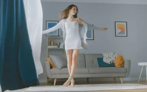 Frau tanzt im Wohnzimmer, die Gardinen wehen. Sie fragt sich, wie oft Gardinen waschen, um die Frische zu bewahren.