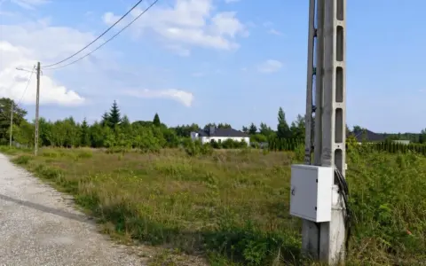 Betonowe słupy energetyczne stoją na tle zieleni i nieba. W oddali widać domki jednorodzinne.