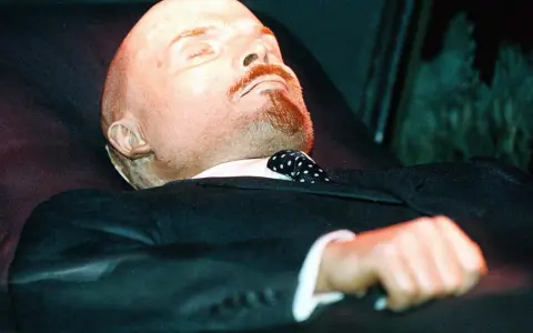 Mumia Lenina w garniturze i z krawatem w kropki, spoczywająca w szklanej gablocie.