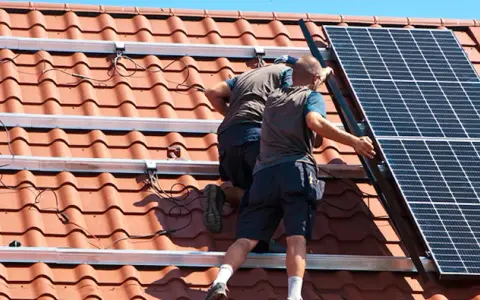 Två personer arbetar med att montera solceller på ett tegeltak. De fäster solpanelerna på monteringsskenor.