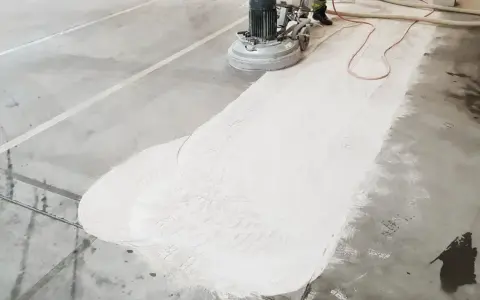 Maszyna szlifuje betonową posadzkę, przygotowując ją do naprawy pęknięć. Biały pył pokrywa część podłogi.