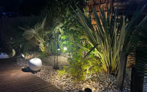 Jardin illuminé la nuit, créant une ambiance chaleureuse. La **norme éclairage extérieur** est respectée pour mettre en valeur la végétation luxuriante et le chemin en bois.
