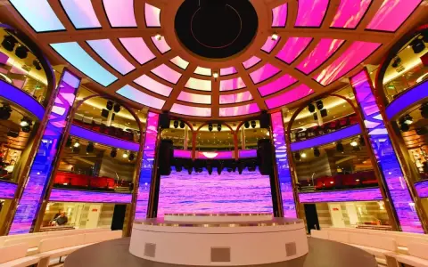 Interni di una nave da crociera, con un palco illuminato da luci rosa e blu, che ricorda la costa mediterranea.