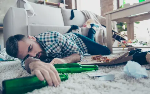 Mężczyzna śpi na dywanie, otoczony pustymi butelkami i resztkami jedzenia. To obrazek pokazujący skutki nadużywania alkoholu, jak padaczka alkoholowa po jakim czasie picia może się pojawić.