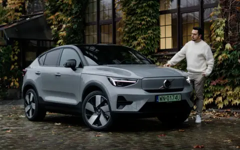 Szary Volvo C40 z czarnym dachem stoi na mokrej, brukowanej nawierzchni. Mężczyzna w białym swetrze opiera się o maskę.
