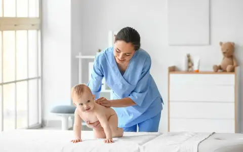 Uśmiechnięty niemowlak na białym podłożu, a nad nim pochylona fizjoterapeutka dla niemowląt w niebieskim uniformie delikatnie masuje jego plecy.