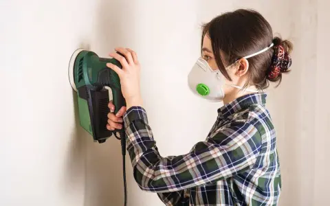Mujer con mascarilla lijando una pared con una lijadora eléctrica.