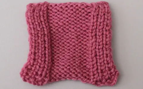 En stickad provlapp i rosa garn, med en vriden maska längs kanterna som ger en vågig effekt.