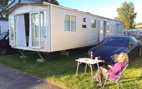 En person njuter av solen vid ett campingbord utanför en husvagn. En Tesla parkerad bredvid. Camping Timrå.