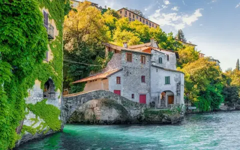 Antica casa in pietra con edera su un lago turchese, un ponte ad arco e colline verdi sullo sfondo, un angolo da fiaba dei castelli Lombardia.