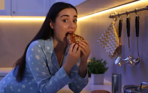Kobieta w piżamie z gwiazdkami je kanapkę, choć czuje, że jest głodna, ale nie ma apetytu. Na blacie leży też pizza i burger.