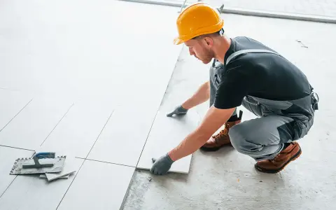Fachowiec w żółtym kasku układa białe płytki. Zastanawiasz się, ile kosztuje położenie kafelek?