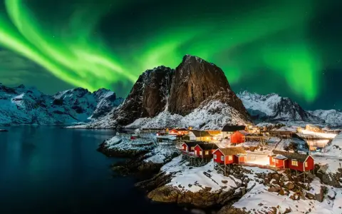 Grön norrsken dansar över snöklädda berg och röda fiskestugor vid en vik i Norge.