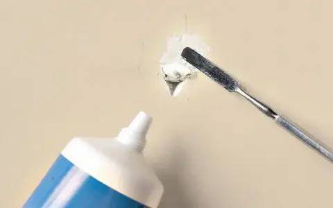 Naprawianie małej dziury w ścianie za pomocą szpachlówki z tubki i metalowej szpatułki.