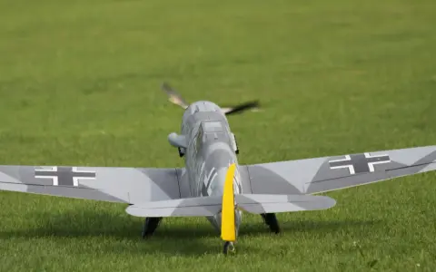 Modellflugzeug einer Me109 mit Tarnmuster und gelbem Seitenleitwerk auf einer grünen Wiese.