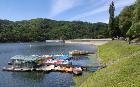 Jezioro Rożnowskie zaprasza na wodną trasę. Na pomoście czekają kolorowe rowerki wodne i łodzie, a w tle wznosi się imponująca zapora.