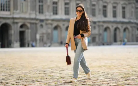 Frau in weiter Jeans und blauem Hemd mit goldener Clutch und Glitzer-Schuhen überquert Straße.