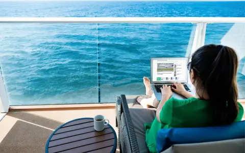 Donna su balcone di nave MSC lavora al laptop, con mare blu sullo sfondo. Scopri il pacchetto WiFi MSC e il suo costo per restare sempre connesso.