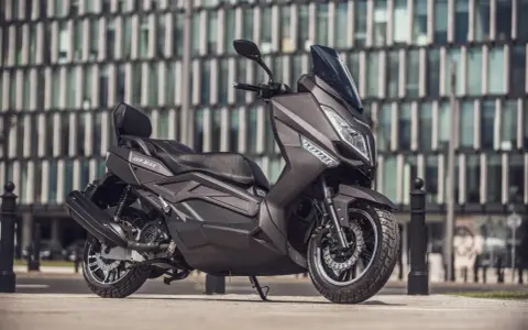 Ciemnoszary skuter Zipp QR Max 125 z czarną tapicerką i czerwonymi przeszyciami, zaparkowany przed nowoczesnym budynkiem.