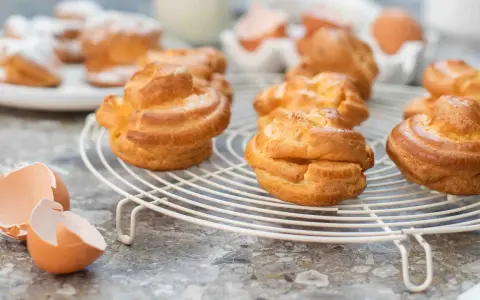 Gyllene petit choux på ett vitt galler, med äggskal och ägg i bakgrunden.
