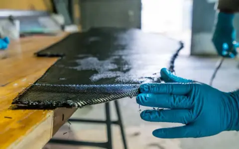 Main d'une personne en gant bleu manipulant un tissu de carbone imbibé de résine, une étape clé pour comprendre qu'est-ce que l'epoxy dans la fabrication composite.