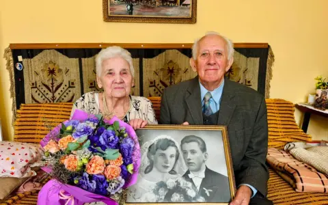 Para świętuje 70 rocznicę ślubu, trzymając zdjęcie z dnia zaślubin. W bukiecie kwiatów dominują fiolety i pomarańcze.