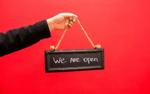 Mano con chaqueta negra sostiene un cartel que dice "We are open", indicando la apertura de un negocio, similar a los tipos de licencia de actividad.
