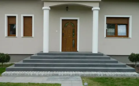 Eleganckie, szare schody zewnętrzne prowadzące do drewnianych drzwi. Świetny pomysł na schody zewnętrzne, podkreślający nowoczesny styl domu.